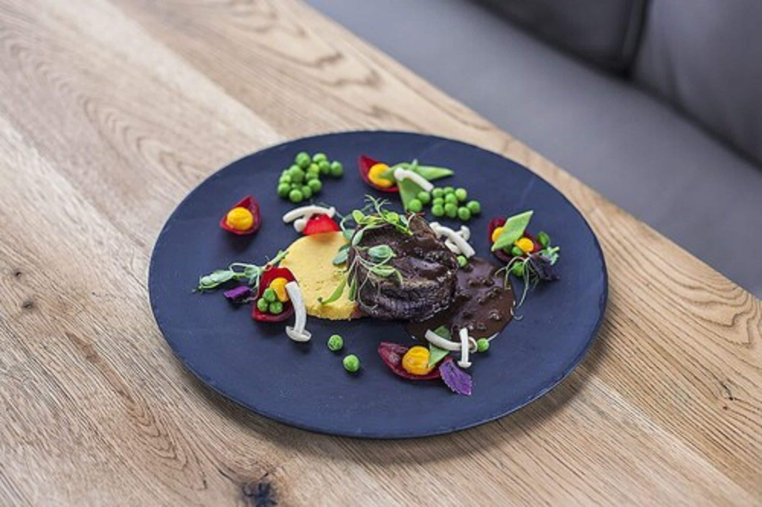 Een rond bord met steak, gepureerde aardappelen en diverse kleurrijke groenten op een houten tafel.