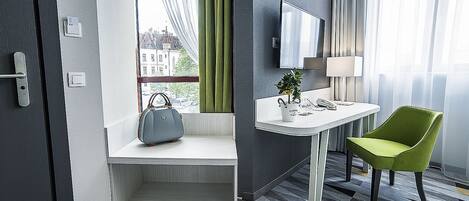 Habitación de hotel con una banca, un escritorio, una silla, un televisor y un ventanal con cortinas.