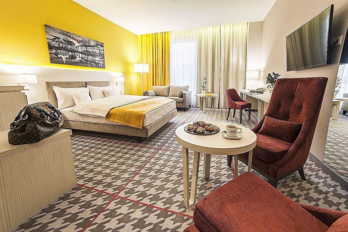 A hotel room with a bed, a round table with a plate of pastries, a red armchair, and a television mounted on the wall.