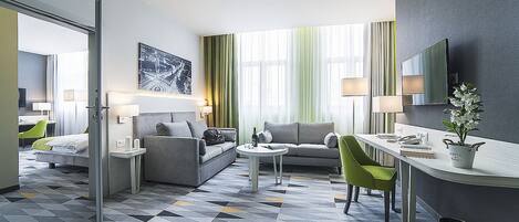 A modern hotel room with a sofa, armchair, and a small dining table. There is a large window with curtains, a flat-screen TV, and a potted plant.