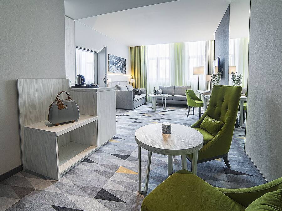 Una habitación de hotel moderna con una zona de estar, un sofá, un sillón verde, una mesita y un bolso decorativo en una estantería.