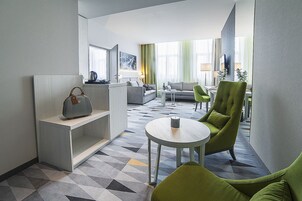 Una habitación de hotel moderna con una zona de estar, un sofá, un sillón verde, una mesita y un bolso decorativo en una estantería.
