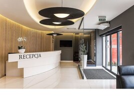 Un área de recepción moderna con un mostrador blanco curvo identificado con la etiqueta “RECEPTION”, una gran lámpara colgante negra y una planta en maceta.