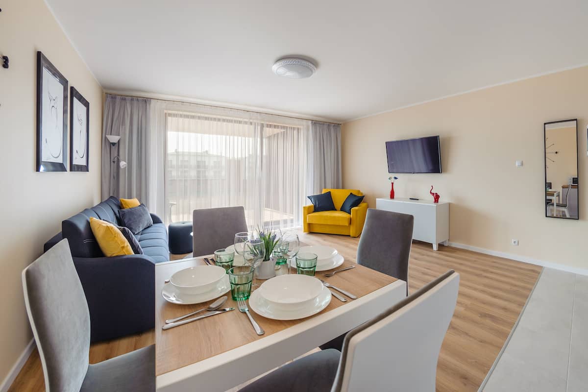 Ein modernes Wohnzimmer mit Esstisch für vier Personen, einer Couch, einem gelben Sessel und einem flachbildfernseher an der Wand.