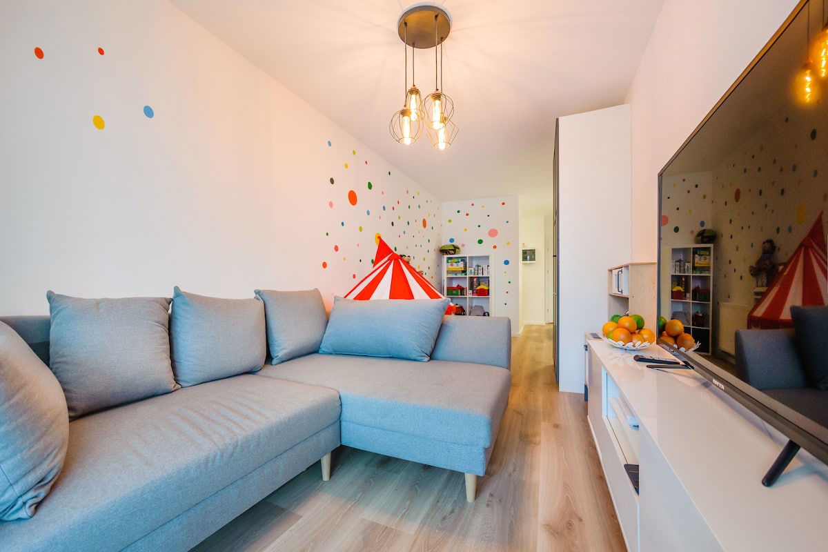 A modern living room with a sectional sofa, a dining area with a white table, and a kitchen with a large mirror.