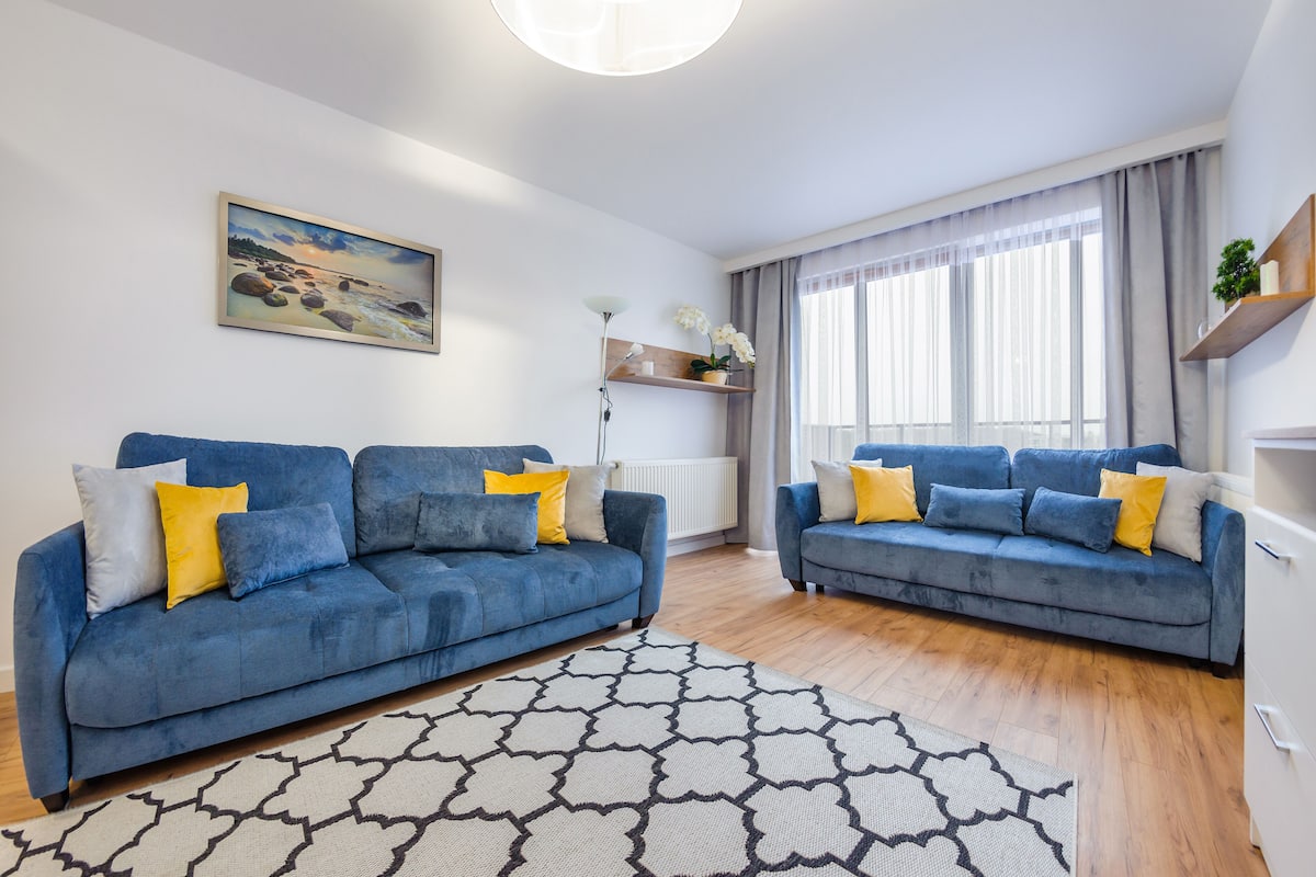 A modern living room with two blue sofas, a patterned rug, a painting on the wall, and a window with curtains.