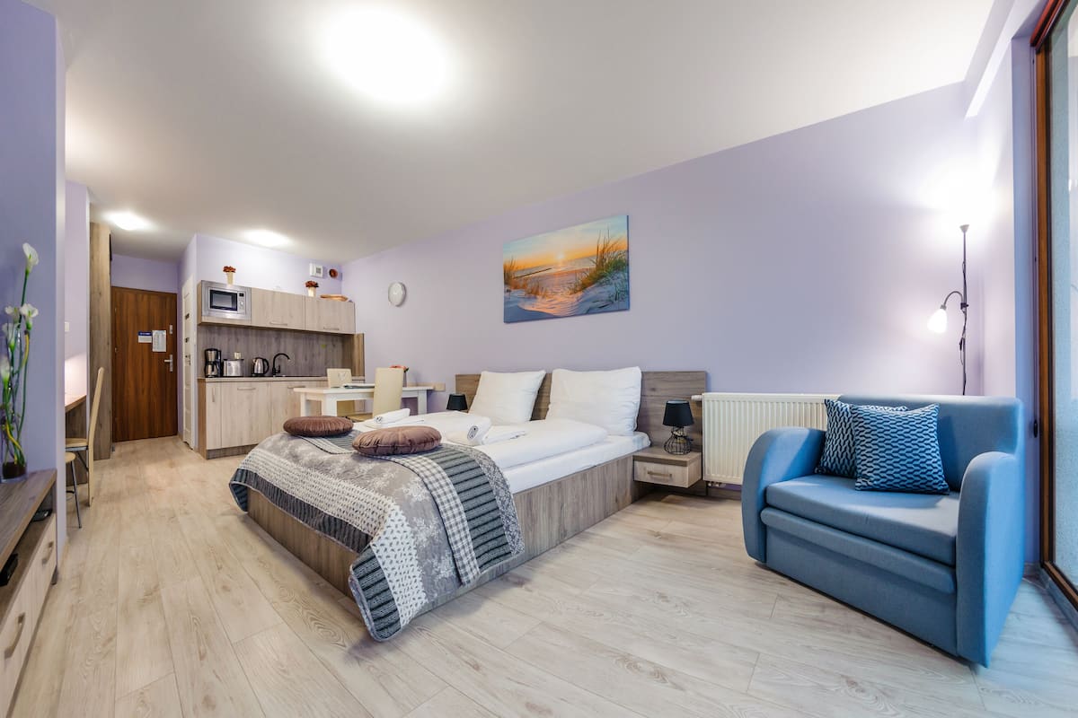 A modern bedroom with a bed, a blue sofa, a kitchenette, and a painting on the wall.
