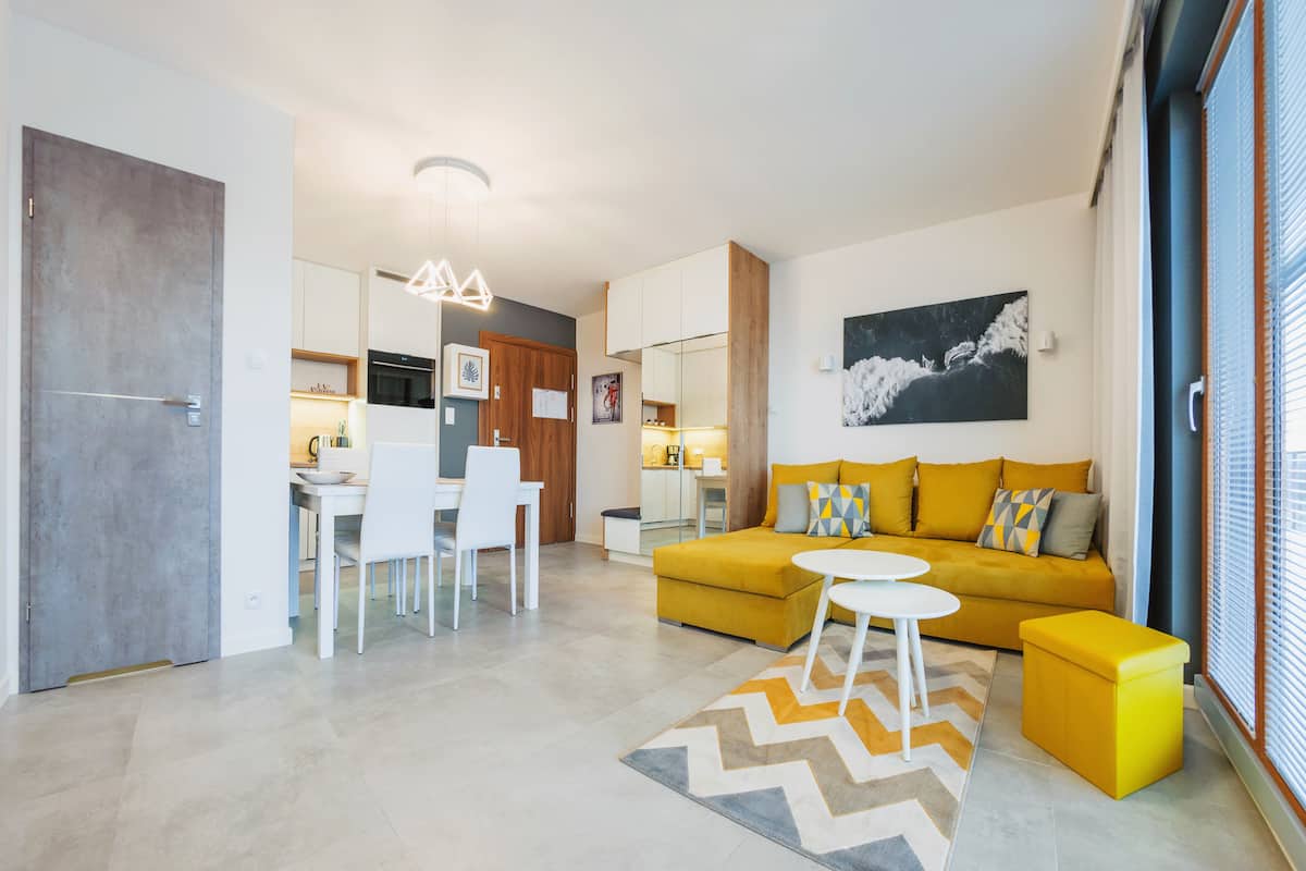 A modern living room with a yellow sofa, a dining area with white chairs, and a kitchen in the background.