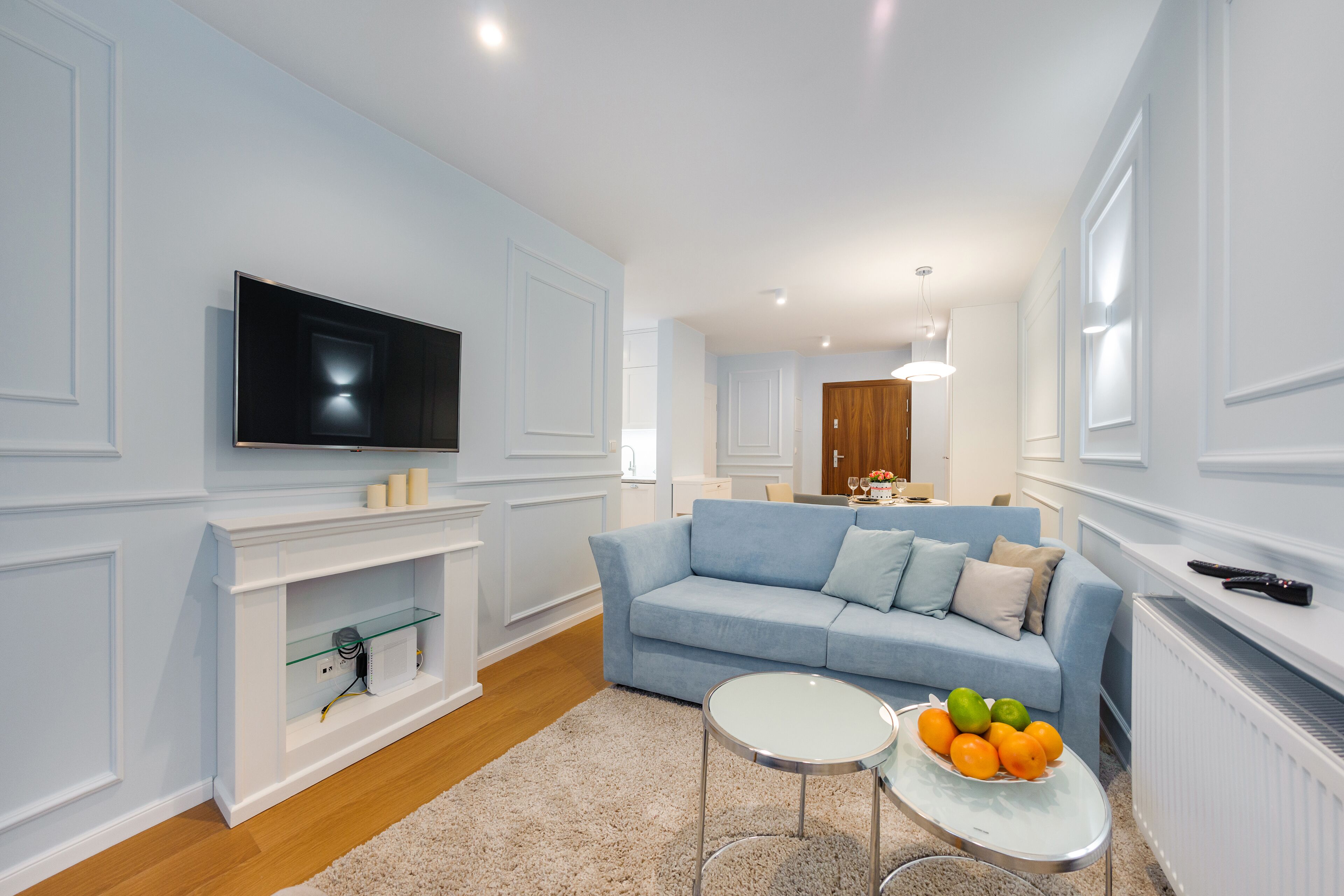 Ein modernes Wohnzimmer mit einem blauen Sofa, einem an der Wand befestigten Flachbildfernseher, einem Glastisch und einem kleinen runden Tisch mit Obst.