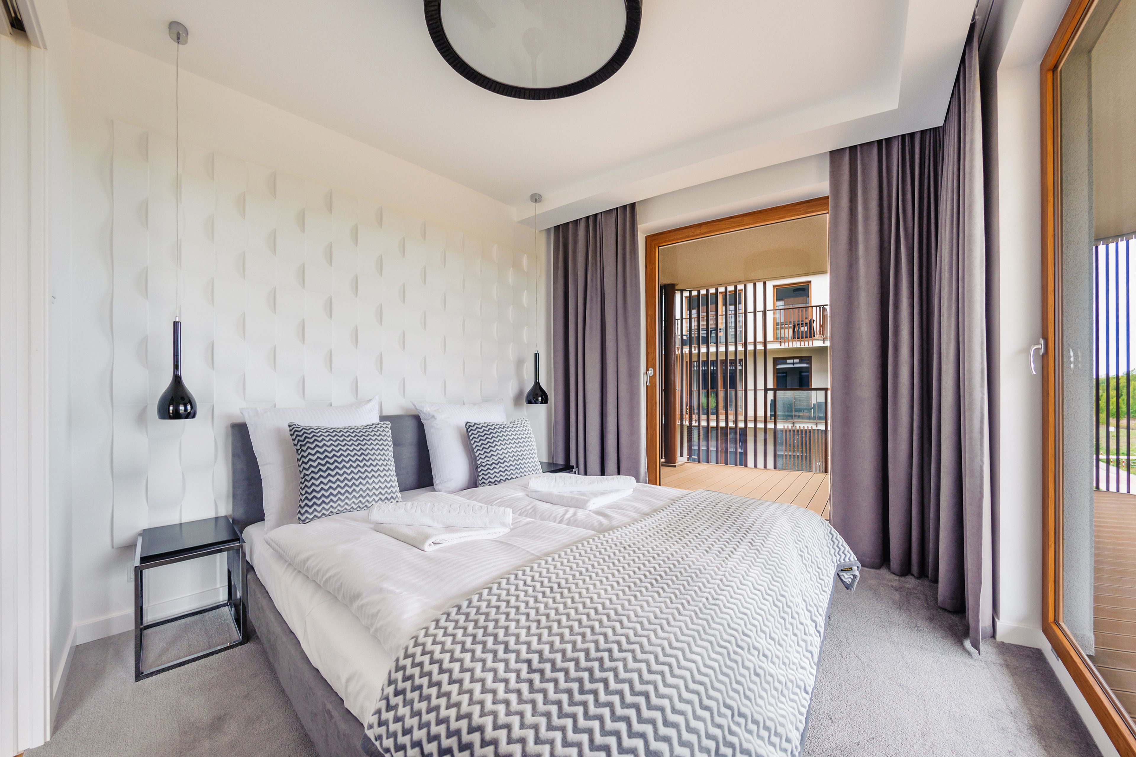 Una habitación de hotel moderna con una cama grande, una mesita de noche, una lámpara de pared y vistas al balcón exterior.