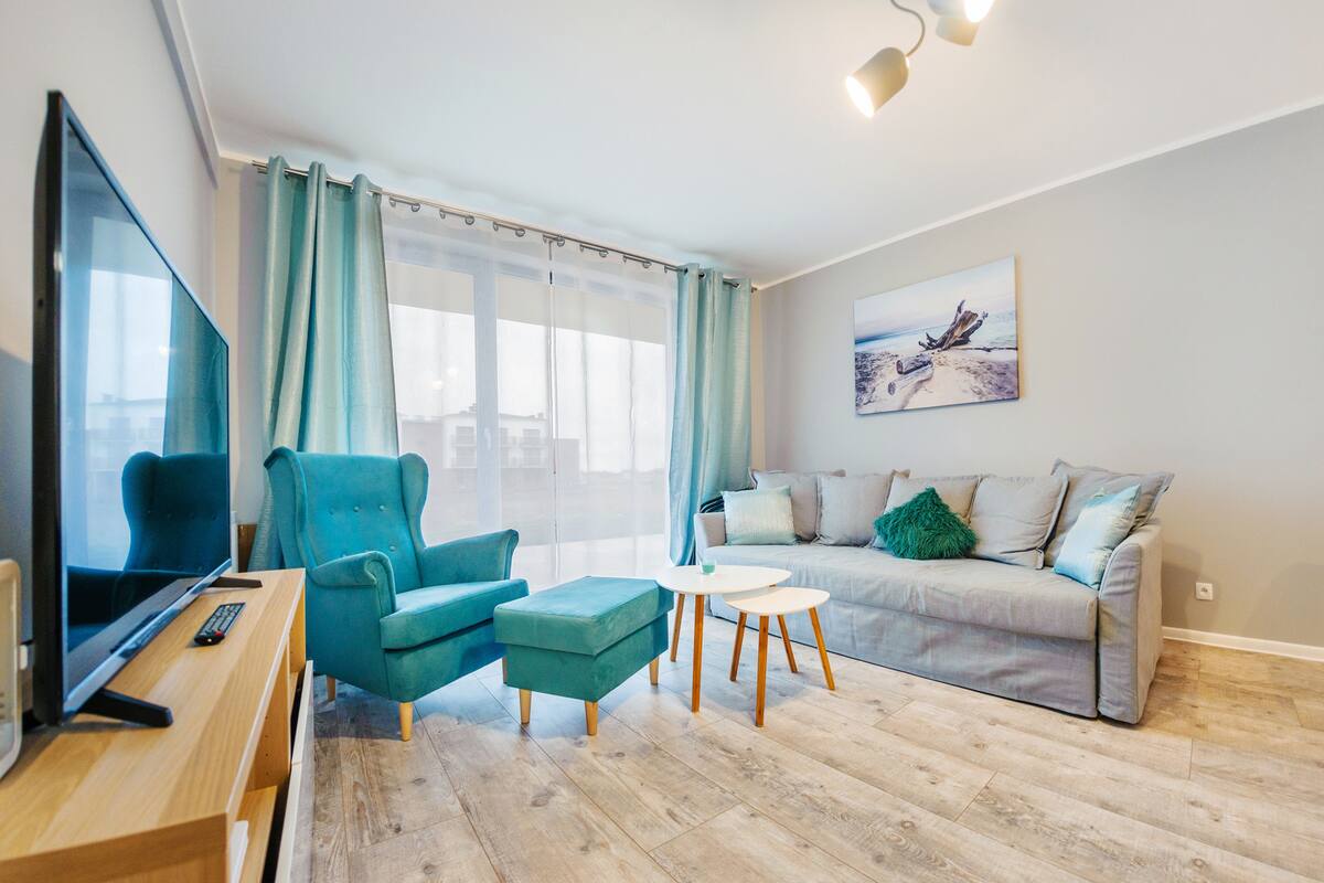 A modern living room with a TV, teal armchair, ottoman, grey sofa, and a painting on the wall.