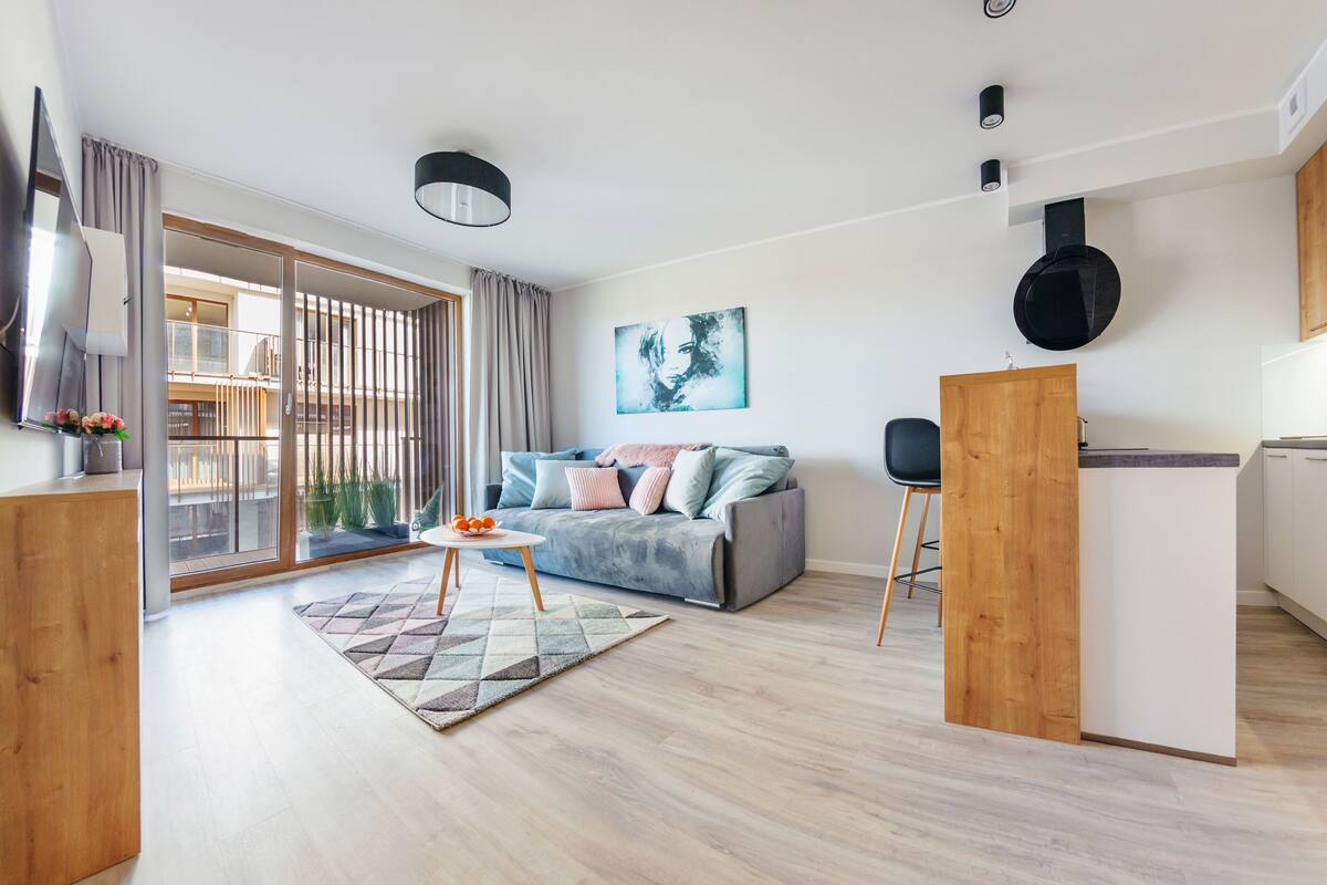 A modern living room with a grey sofa, wooden accents, a kitchen area, and a balcony with large windows.