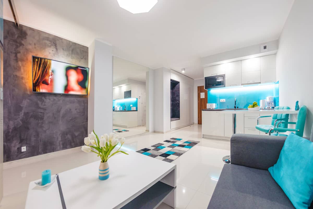 A modern living room with a sofa, a coffee table with a vase of flowers, a kitchen area with a blue countertop, and a large mirror reflecting the room.
