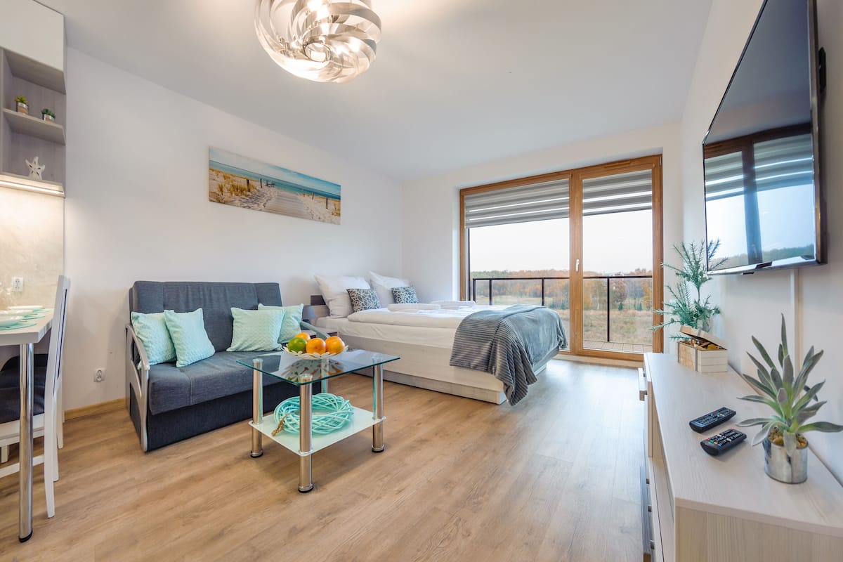 Ein modernes Wohnzimmer mit einer Couch, einem Glastisch mit Früchten, einem Bett mit Blick und einem Fernseher.