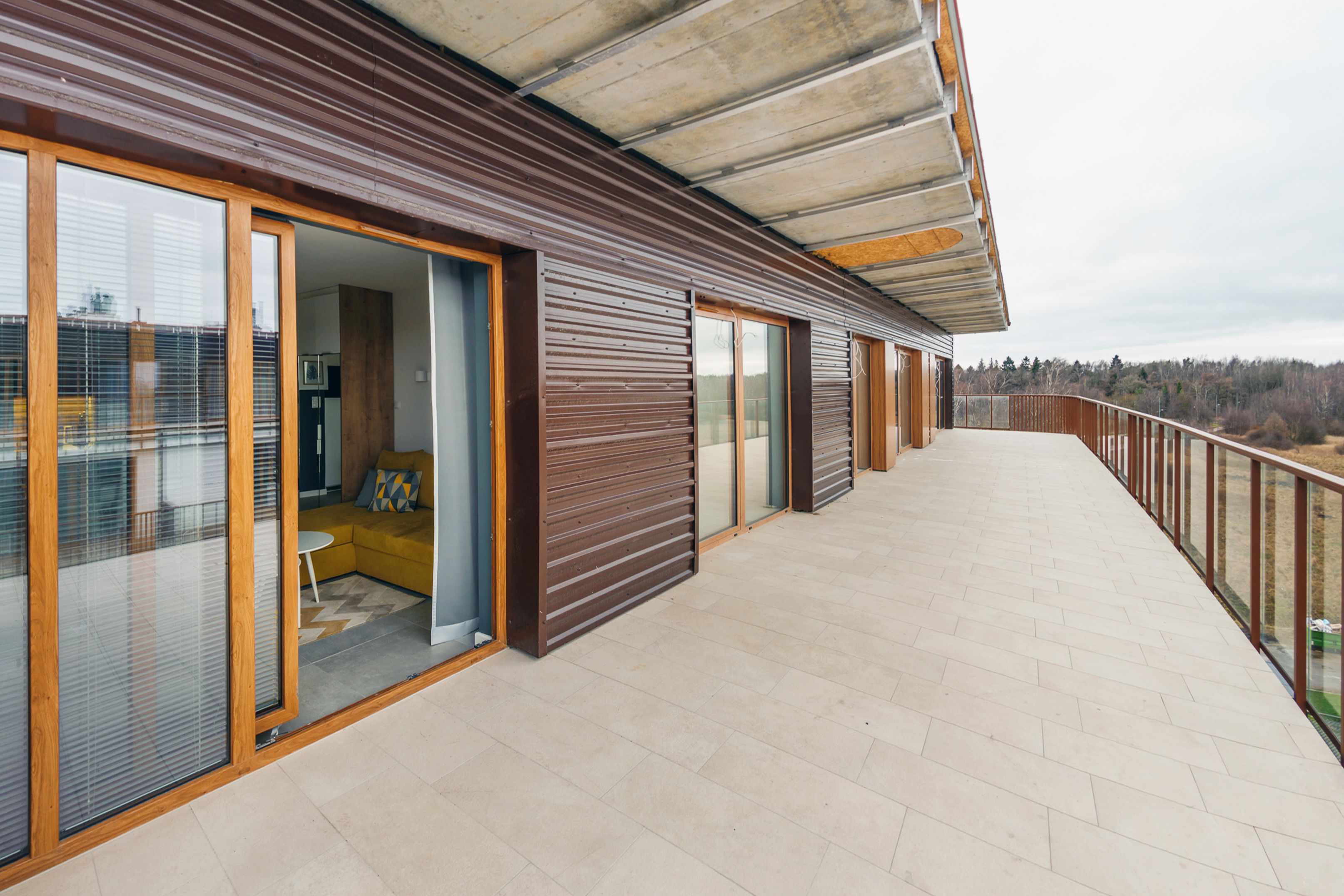 A modern building with large sliding glass doors, a balcony with a metal railing, and a herringbone-patterned floor.