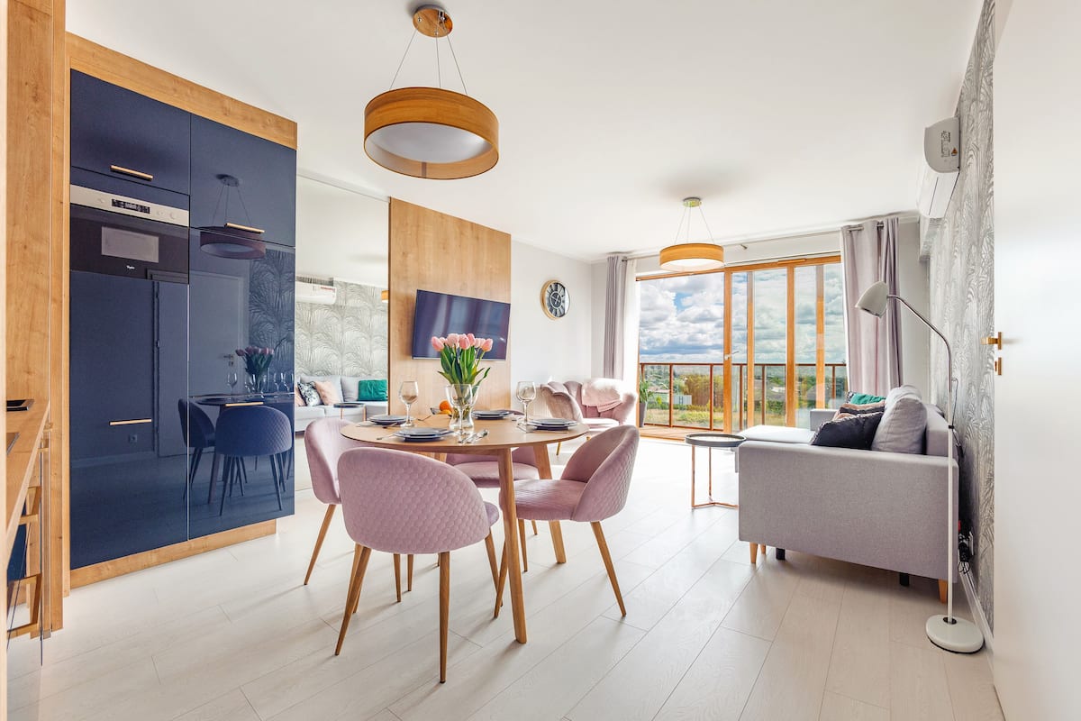 A modern kitchen with built-in appliances, a dining area with pink chairs, and a living room with a grey sofa.