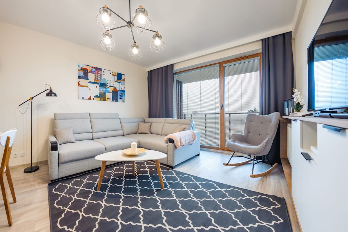A modern living room with a sectional sofa, a rocking chair, a coffee table, and a large window with blinds.
