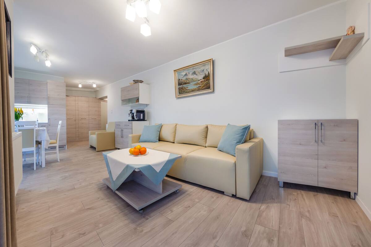 A modern living room with a beige sofa, a dining area with a white table, and a kitchen in the background.