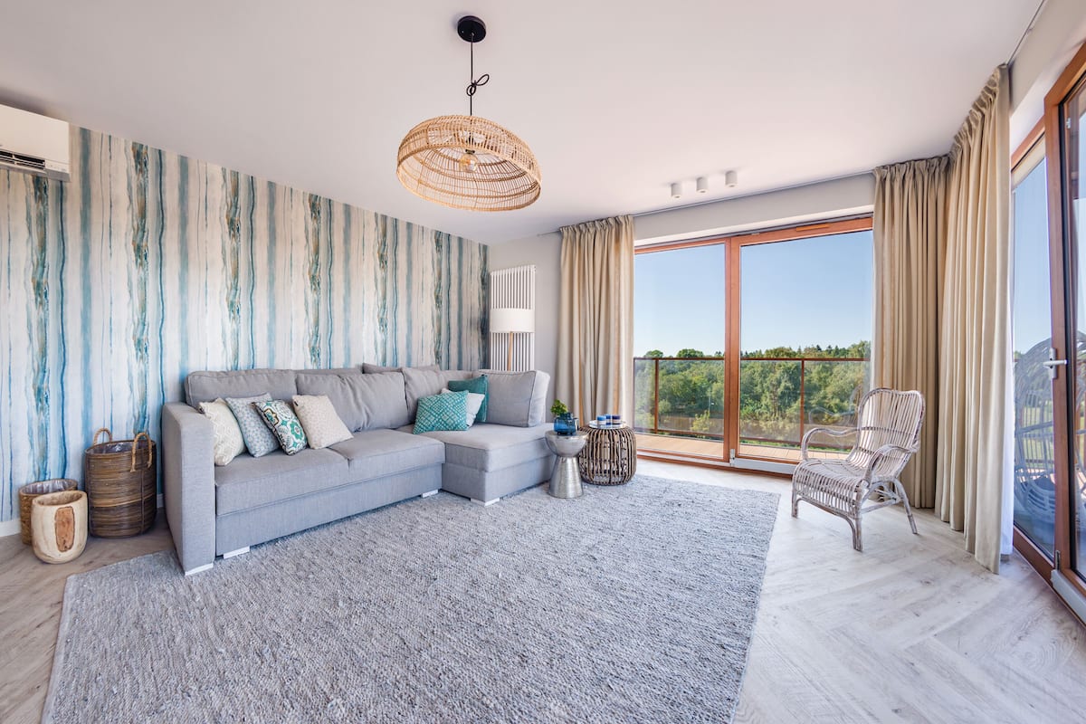 Ein modernes Wohnzimmer mit einem grauen Ecksofa, einem Korbstuhl und einem großen Fenster mit Blick auf Grünflächen.