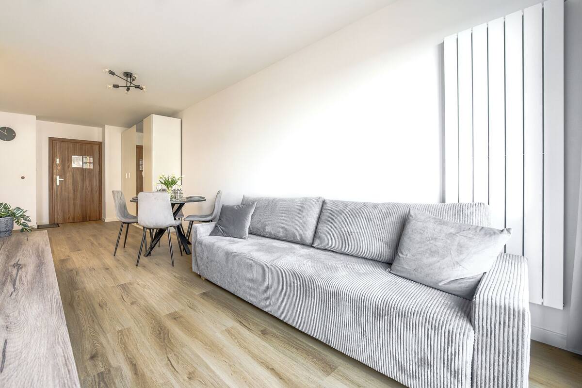 A modern living room with a sofa, dining table, and chairs, wooden flooring, and a ceiling light fixture.