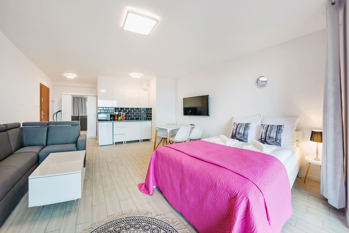 A modern hotel room with a large bed, a sofa, a desk, and a kitchenette.