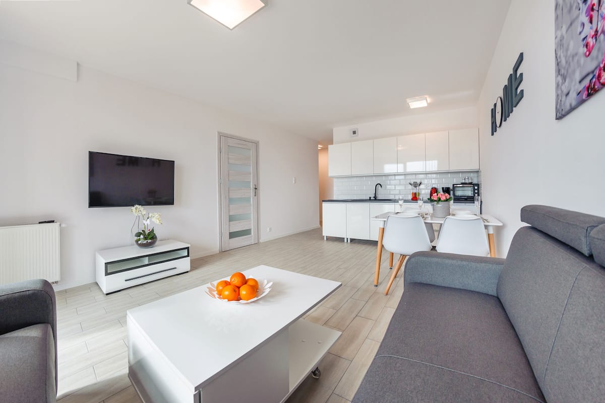 A modern living room with a grey sofa, a white coffee table with oranges, a flat-screen TV, and a kitchen in the background.