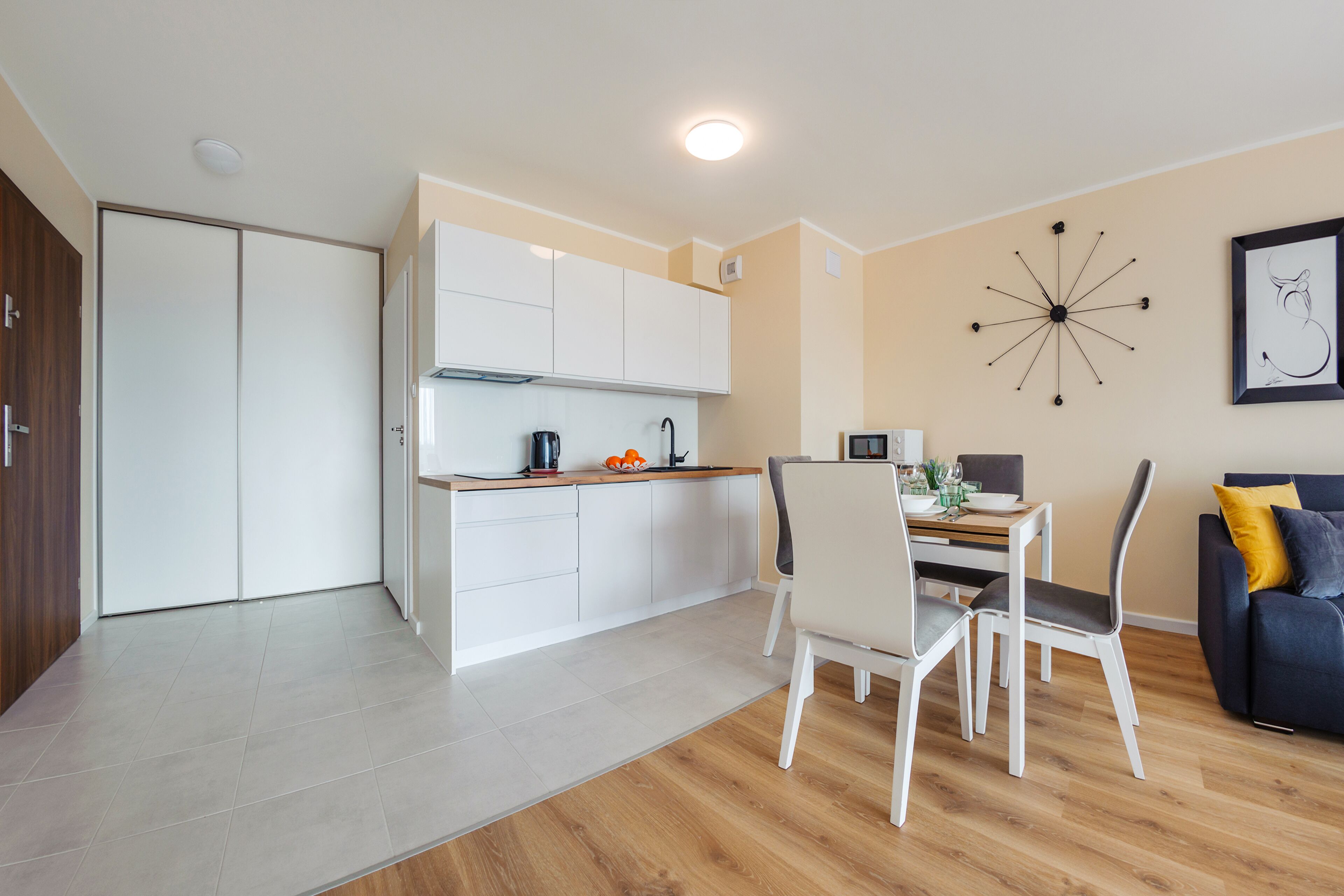 A modern kitchen with white cabinets, a dining area with a table and chairs, and a living area with a sofa and a wall clock.