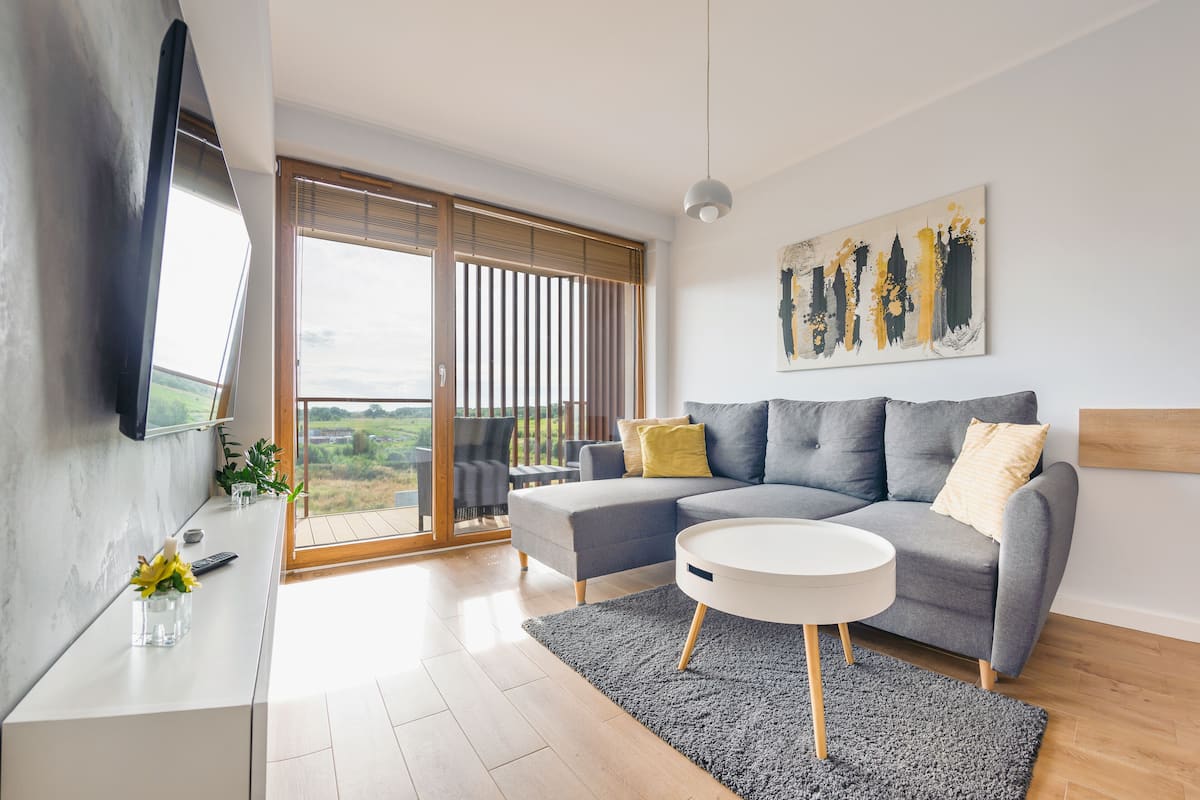 Ein modernes Wohnzimmer mit grauem Sofa, weißem Couchtisch und einem großen Fenster mit Jalousien.