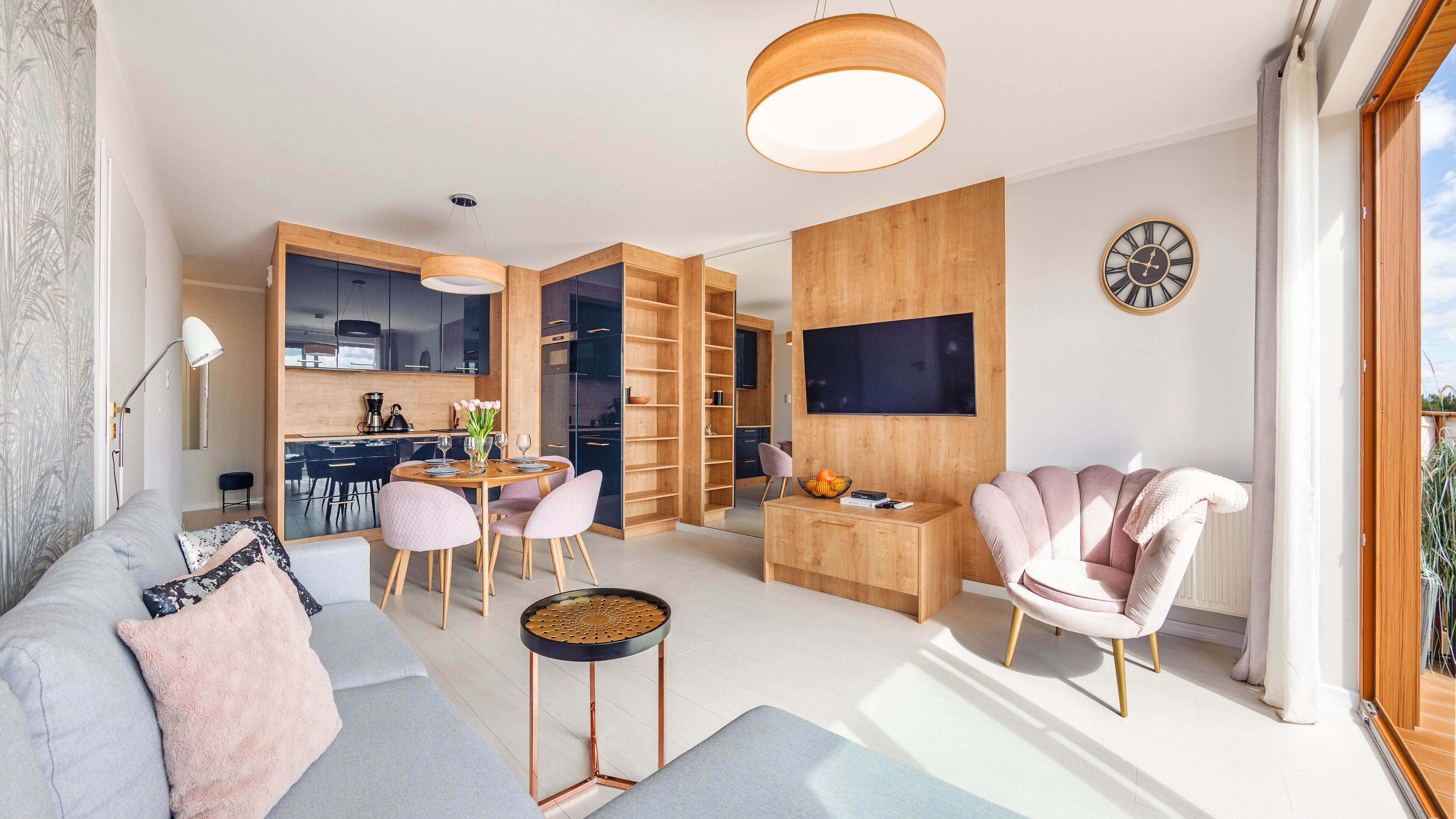 A modern living room with a sofa, a dining table, and a pink armchair. There is a kitchen area with cabinets and a television.