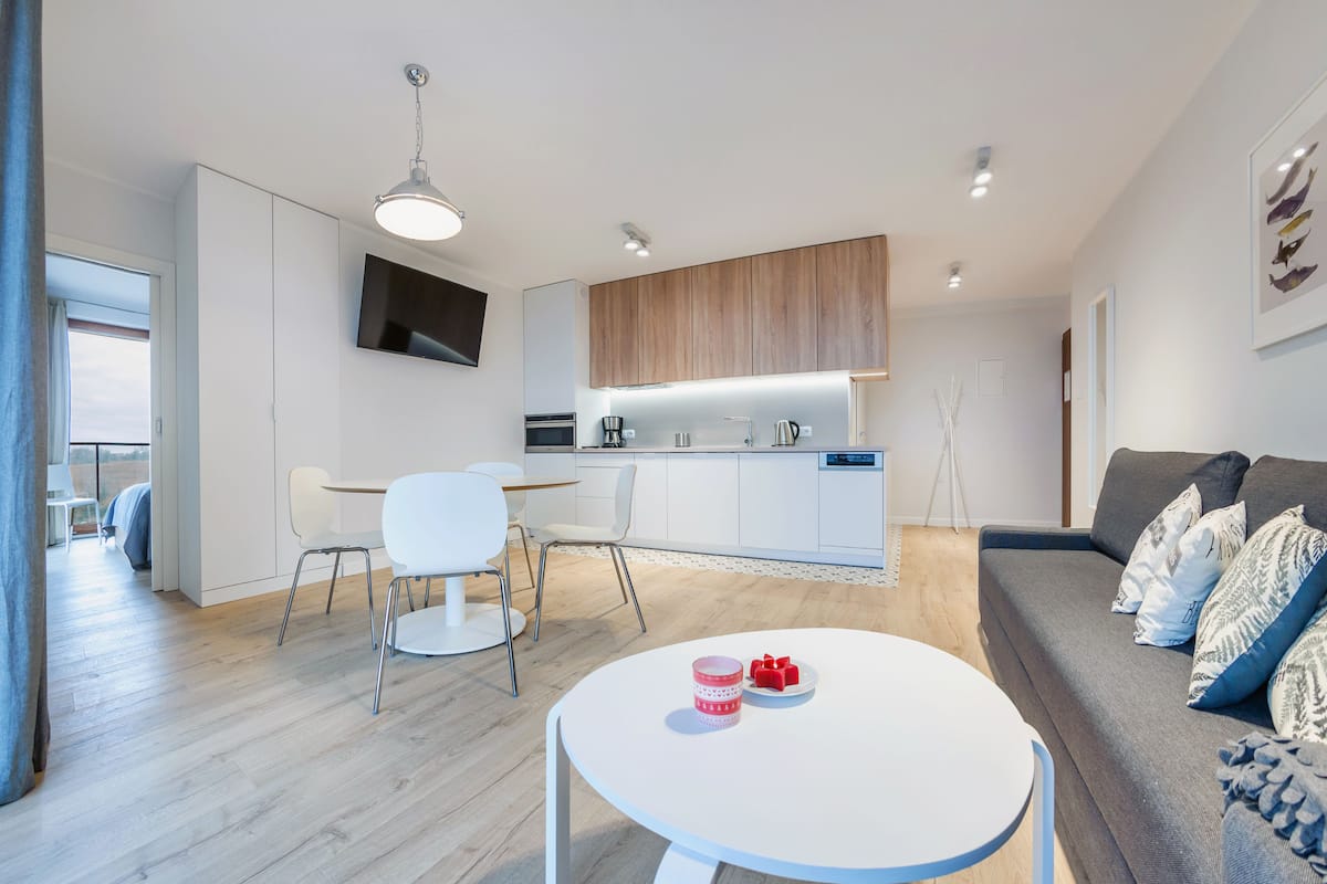A modern living room with a white round table, a grey sofa with patterned cushions, and a kitchen area with white cabinets and a wooden countertop.