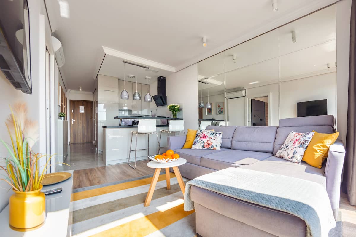 Ein modernes Wohnzimmer mit einer Couch, einem Couchtisch und einem kleinen Beistelltisch. Im Hintergrund ist eine Küchenzeile mit einem Esstisch und Stühlen zu sehen.