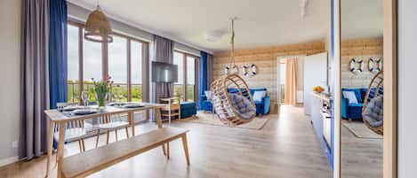 A spacious living area with a dining table, blue sofas, a hanging chair, and a large window offering a view.