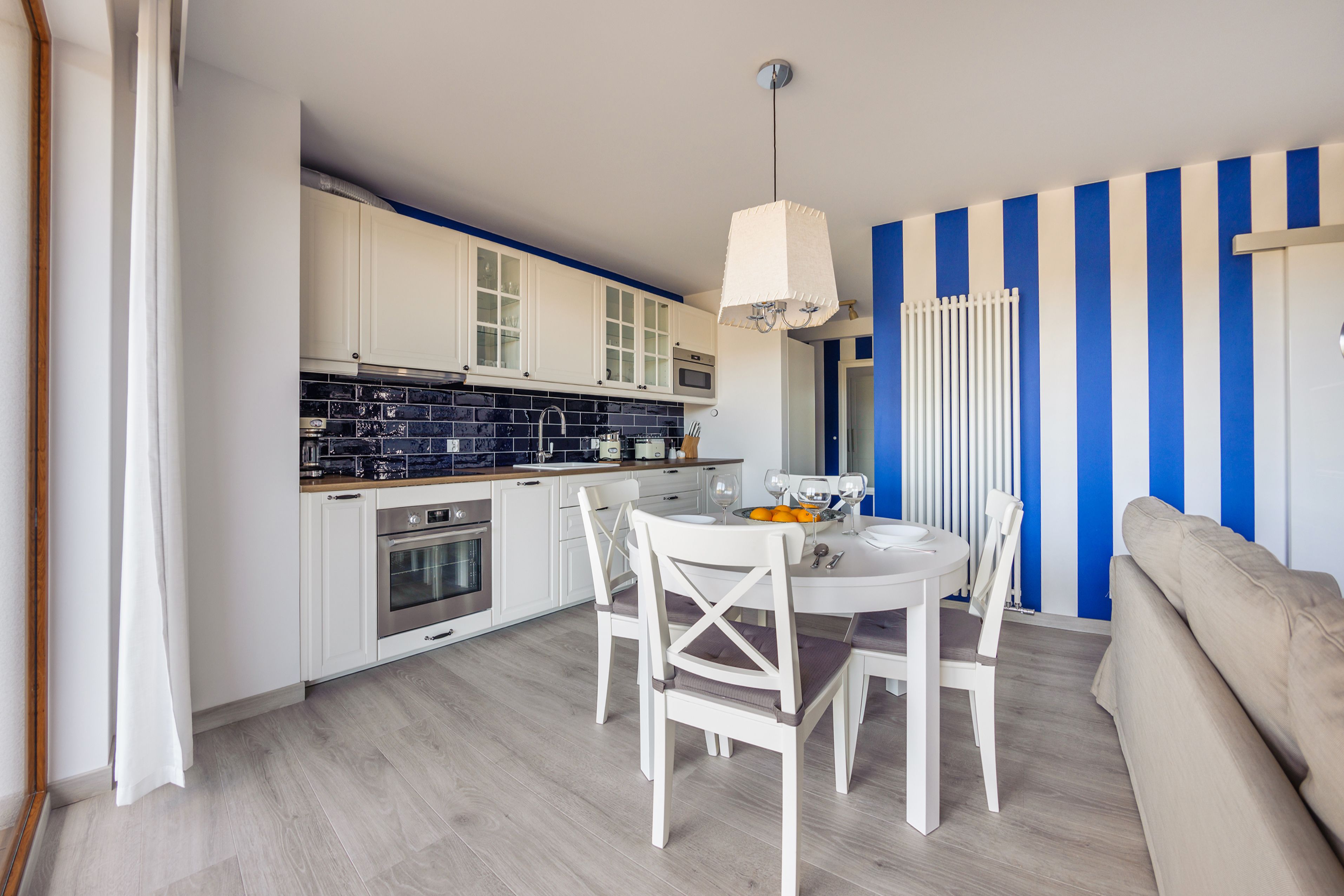 A modern kitchen with white cabinets and a black backsplash, a dining area with a white table and chairs, and a blue and white striped wall.