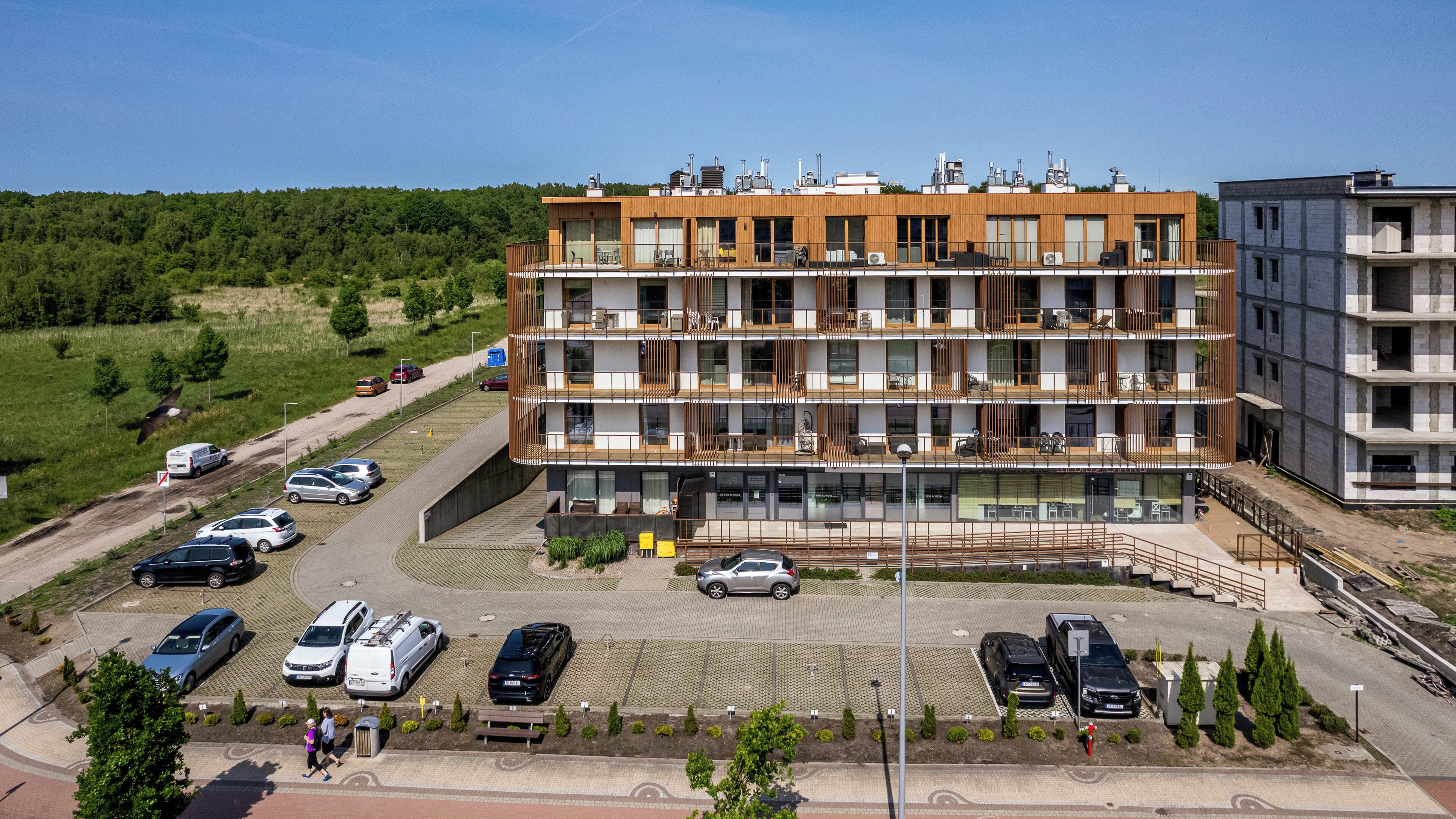 A multi-story residential building with balconies, a parking area, and a surrounding green landscape.