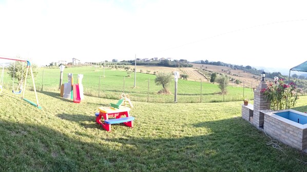 A grassy outdoor area with a swing set, a small play structure, and a brick-built pool with a lamp post and flowers. - Agriturismo Il Vecchio Carro (Campli)