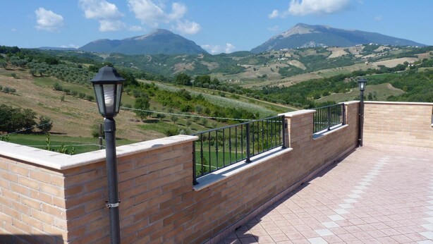 Een terras met een bakstenen muur en metalen hekwerk, met uitzicht op een heuvellandschap met een berg in de verte.