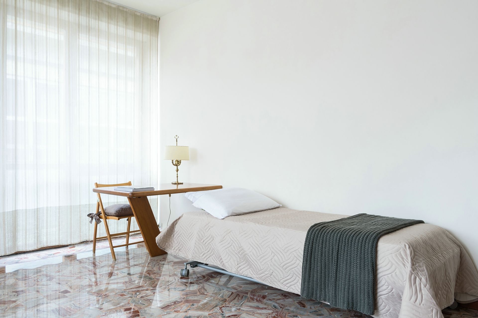 Una cama individual con una colcha, una mesita de noche con una lámpara, una silla y una ventana con cortinas traslúcidas.