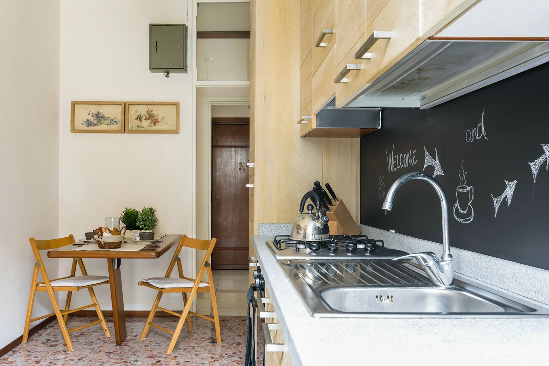 Cocina compacta con estufa a gas, lavamanos y una pizarra con dibujos. Se ve una mesa de madera con sillas y una pequeña planta encima.