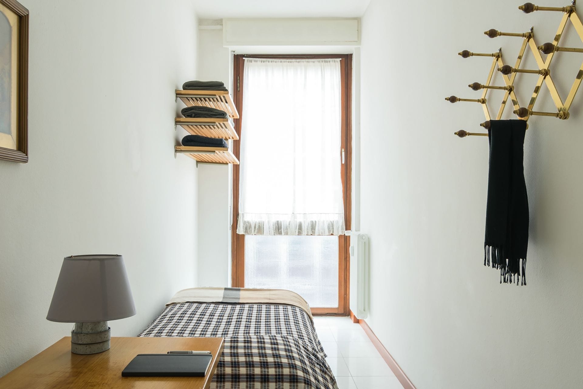 A bedroom with a bed, a bedside table with a lamp, a window with curtains, a wall-mounted drying rack, and a black scarf hanging on a decorative metal structure.