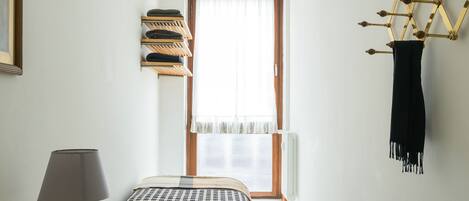 A bedroom with a bed, a bedside table with a lamp, a window with curtains, a wall-mounted drying rack, and a black scarf hanging on a decorative metal structure.
