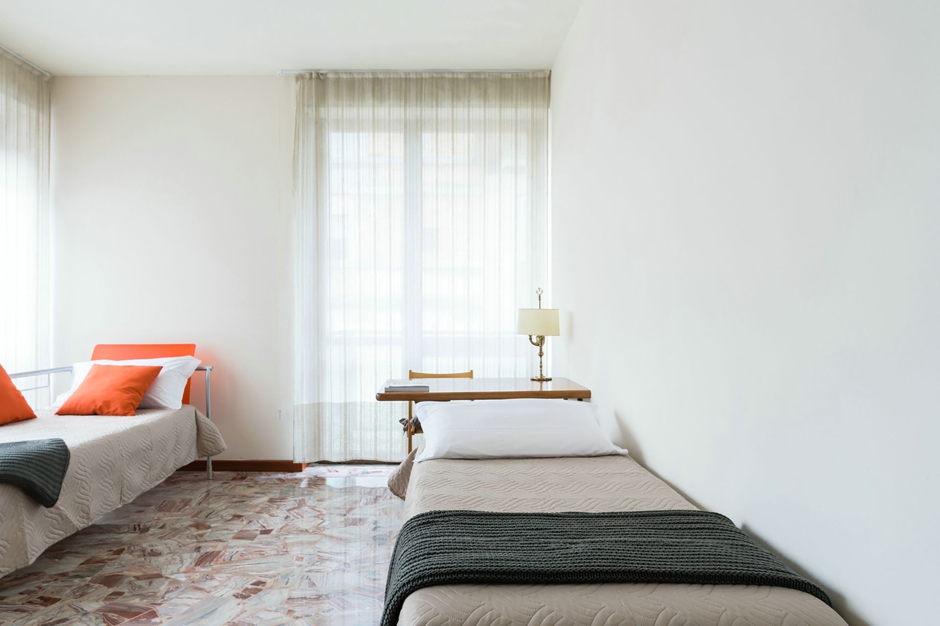 Una habitació d'hotel amb dos llits, una taula petita amb una làmpada i una finestra amb cortines transparents.