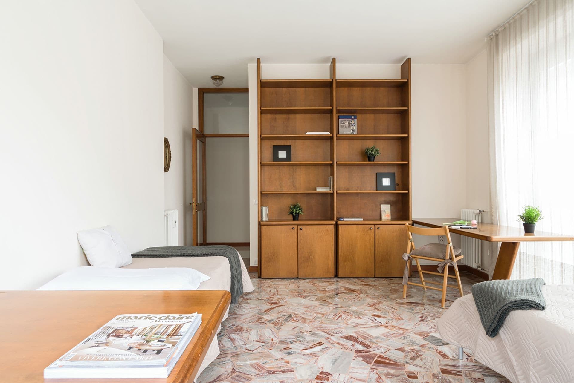 A modern, well-lit room with a bed, a wooden bookshelf, a desk, and a chair.