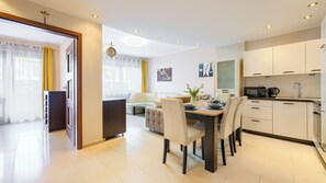 Uma cozinha moderna com armários brancos, um espaço de refeições com mesa posta para quatro pessoas e uma sala de estar com sofá e televisão.