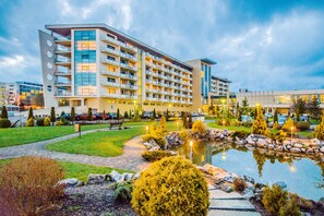 Um hotel moderno com vários andares, um jardim bem cuidado com um lago e uma área de estacionamento.