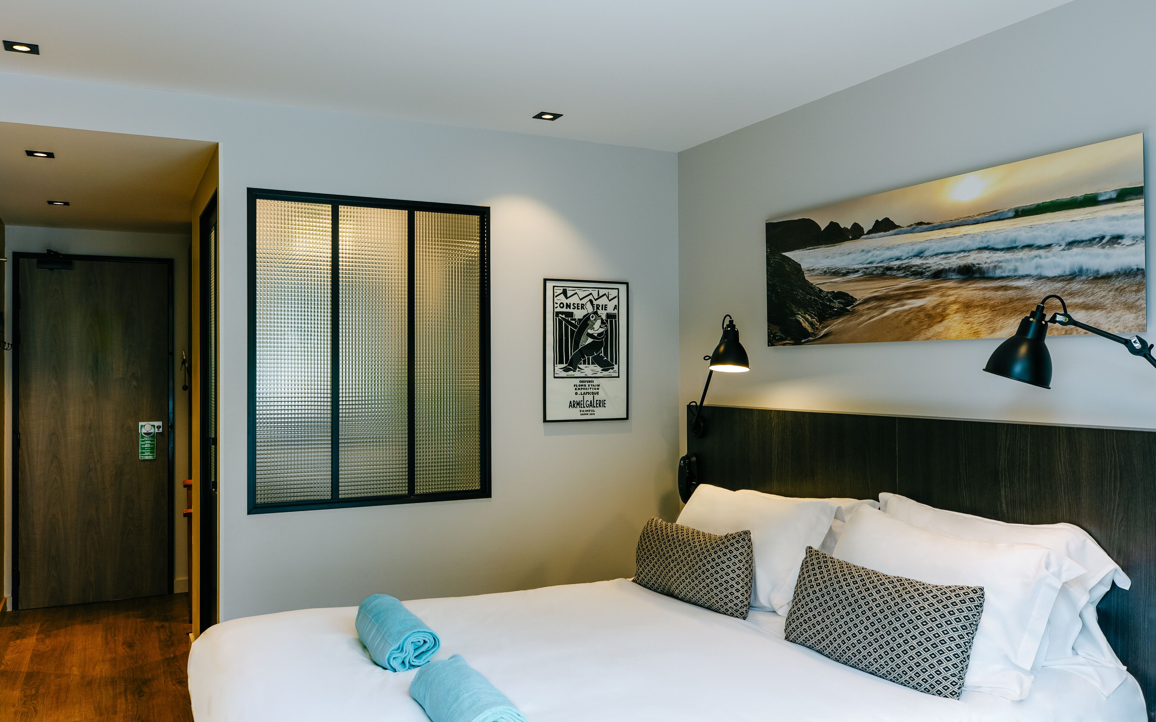 Ein Hotelzimmer mit Bett, einem Fenster mit Jalousien, einem Strandbild und einer Lampe.