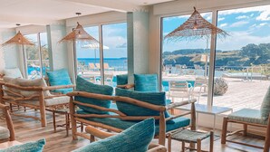A poolside area with wooden lounge chairs and blue cushions, large windows with a view of the sea, and woven ceiling lamps.