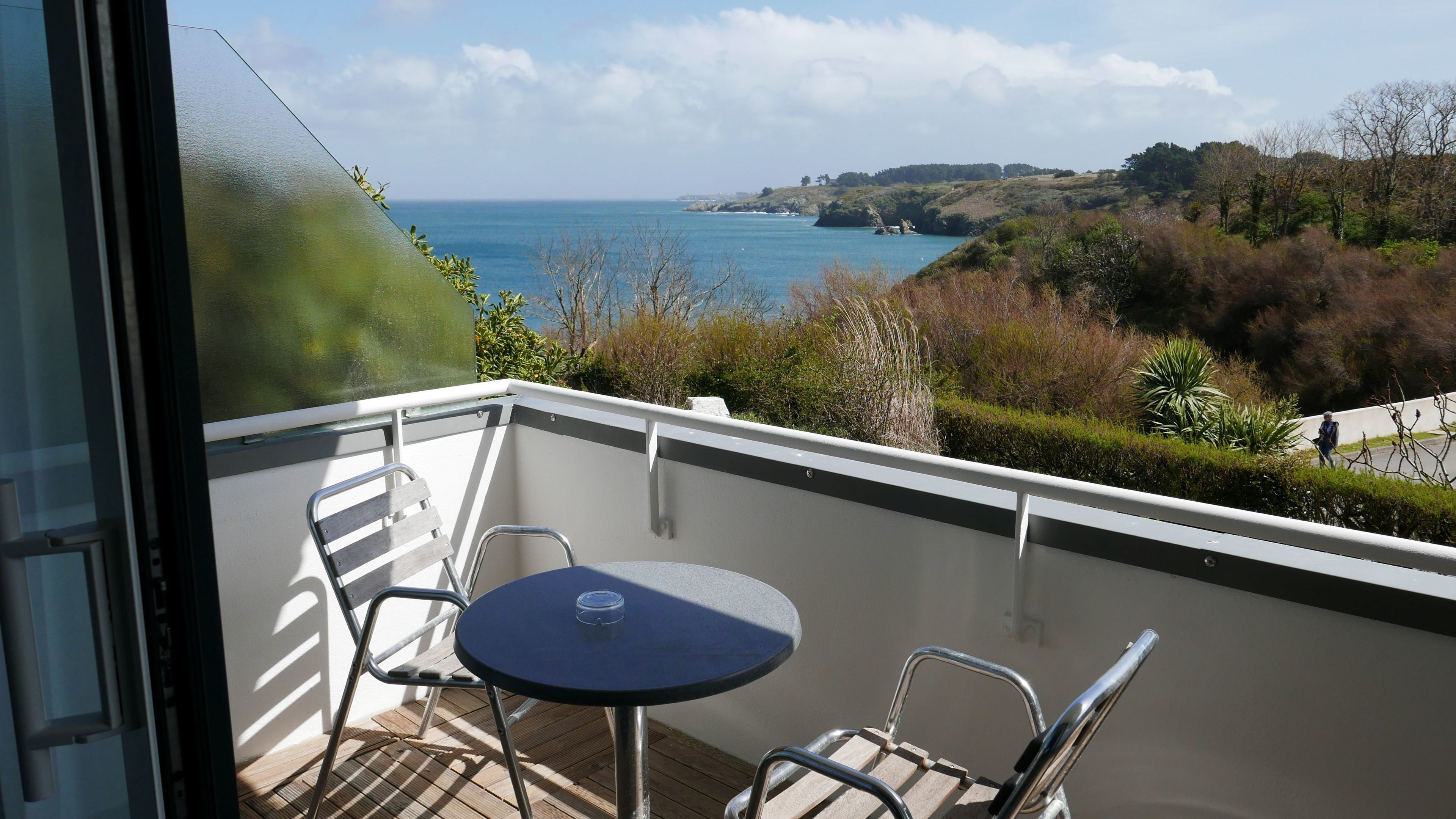 Un balcón con mesa y sillas, con vista a un paisaje costero con colinas y el mar.