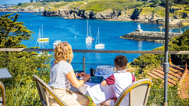 Two people seated at a table with a view of a coastal area, including boats and rocky terrain.