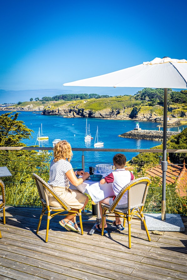 Dos personas sentadas en una mesa con vistas a la costa, incluyendo barcos y terreno rocoso.