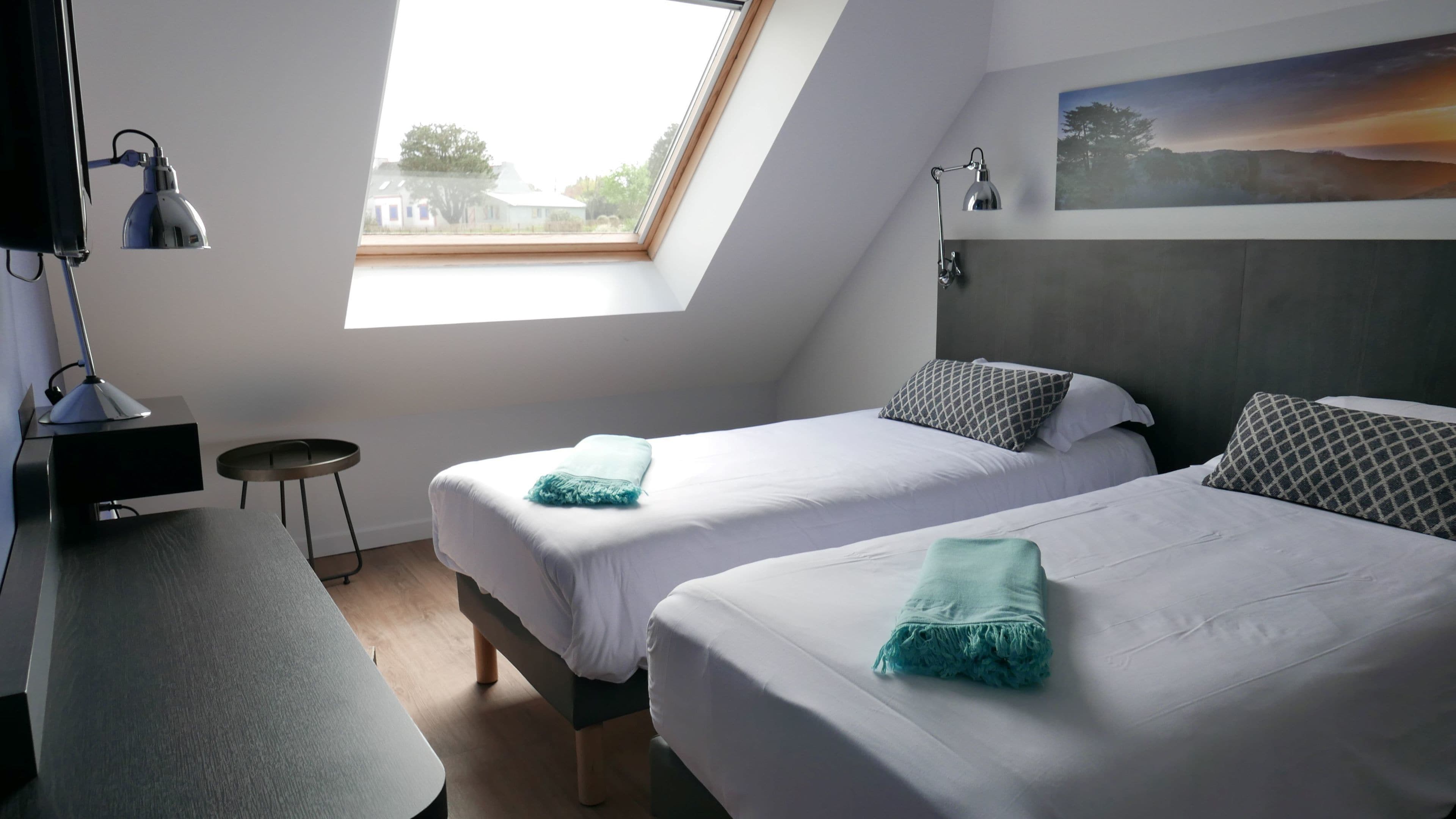 Ett modernt hotellrum med två sängar, ett skrivbord, en stol, ett litet bord och ett takfönster.
